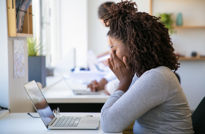 Stressed professional woman sitting at desk with hands covering her face, looking at laptop screen in a bright modern office, showing signs of burnout or mental exhausion.