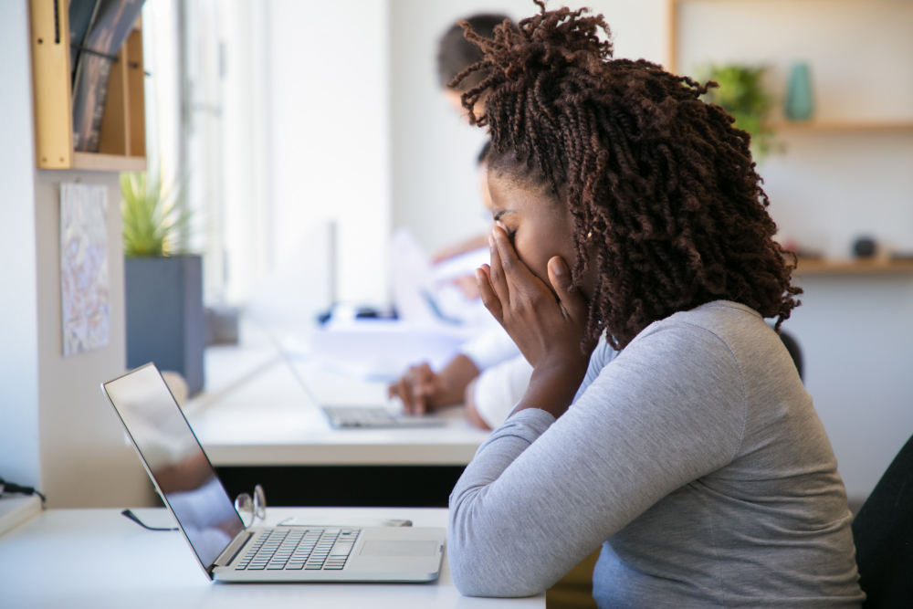 Stressed professional woman sitting at desk with hands covering her face, looking at laptop screen in a bright modern office, showing signs of burnout or mental exhausion.