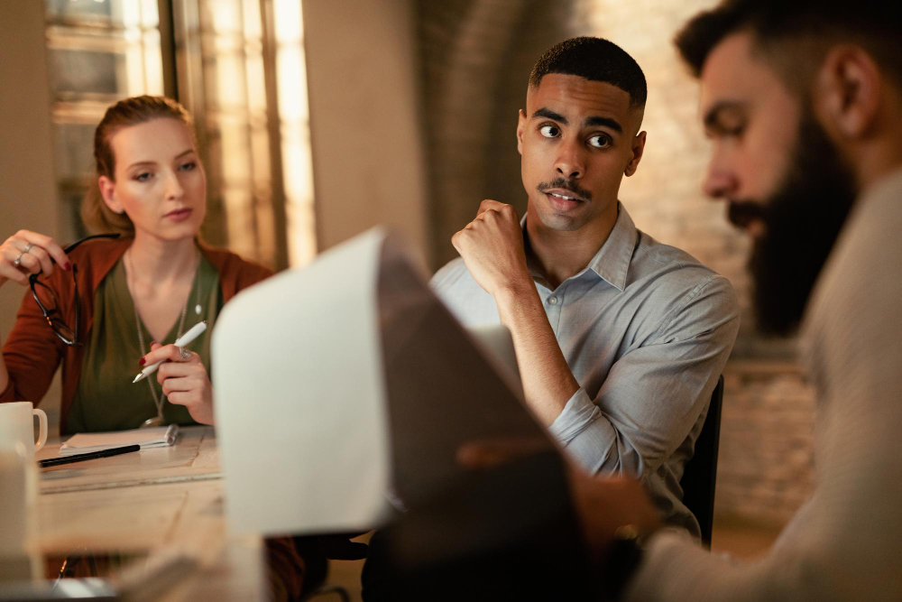 Three colleagues in a serious discussion around a table, reviewing documents in a dimly lit office setting, illustrating open communication and psychological safety during workplace collaboration.