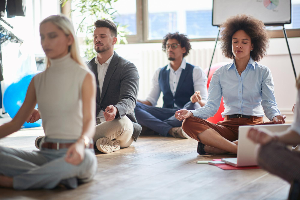 Group of diverse professionals sitting cross-legged on the floor in a bright office space, meditating together with eyes closed during a workplace wellness or mindfulness session.