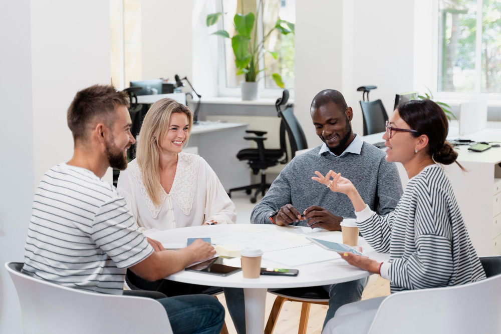 A group of four colleagues sitting around a round table in a bright office, smiling and engaged in conversation, reflecting psychological safety and inclusive team communication.