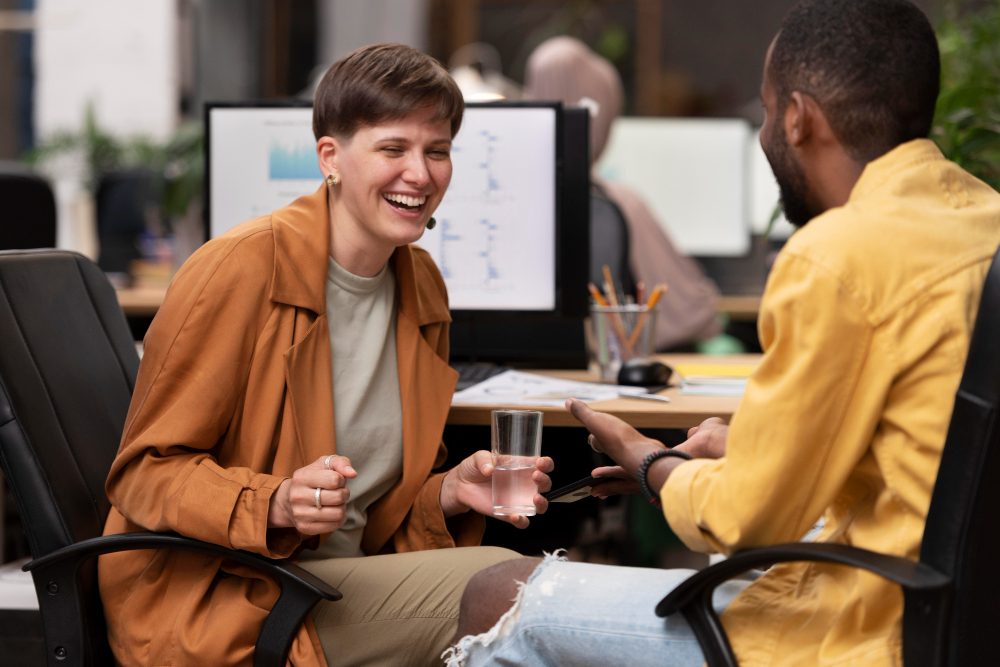 Two colleagues having a friendly, light-hearted conversation in a modern office, with one person holding a glass of water and laughing, suggesting a positive and supportive workplace environment