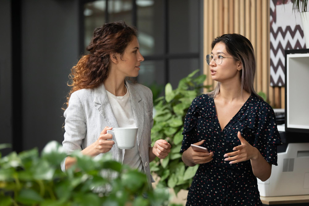 Alt text: Two professional women having a focused conversation in a modern office break area, one holding a coffee mug and the other gesturing while holding a phone, suggesting a collaborative or supportive discussion.