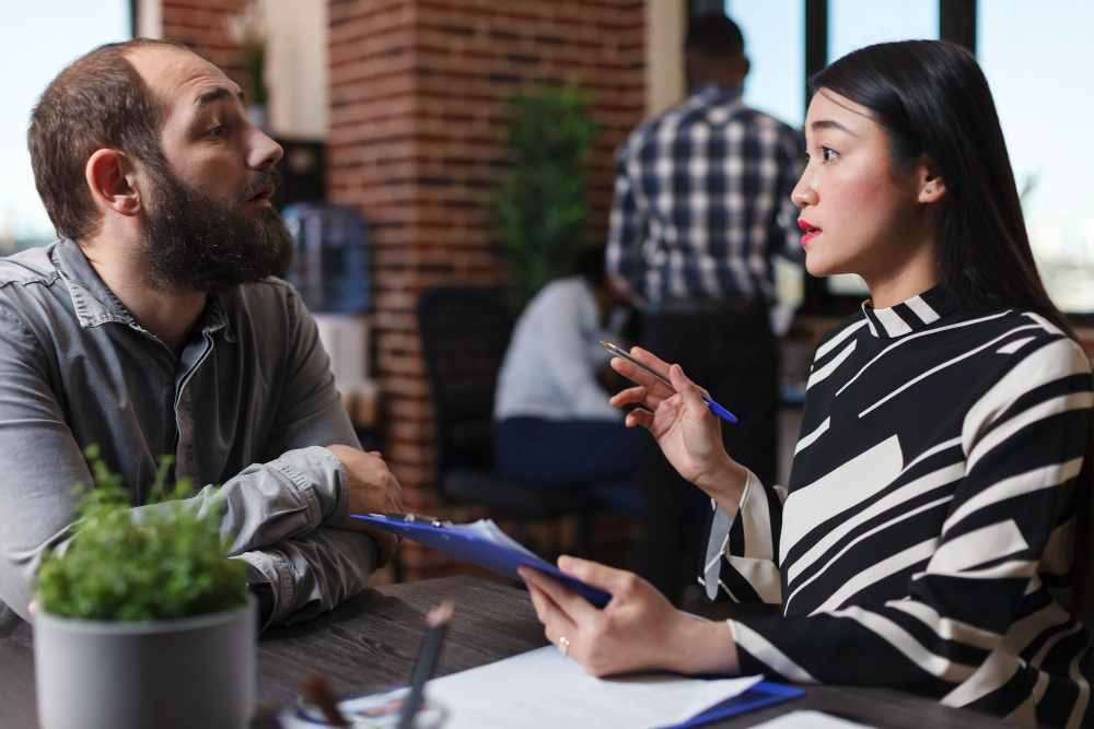 Two coworkers engaged in a thoughtful conversation at a shared desk, with one holding a clipboard, illustrating open communication and psychological safety in the workplace.