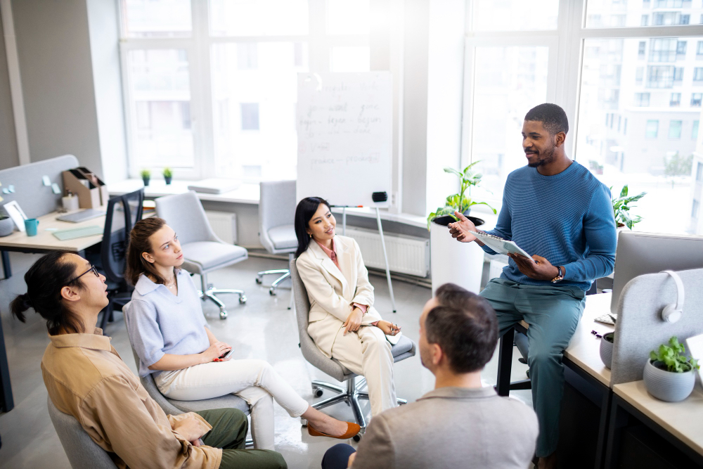 A standing trainer holds a tablet and speaks to a diverse, seated group of employees about shared execution standards.